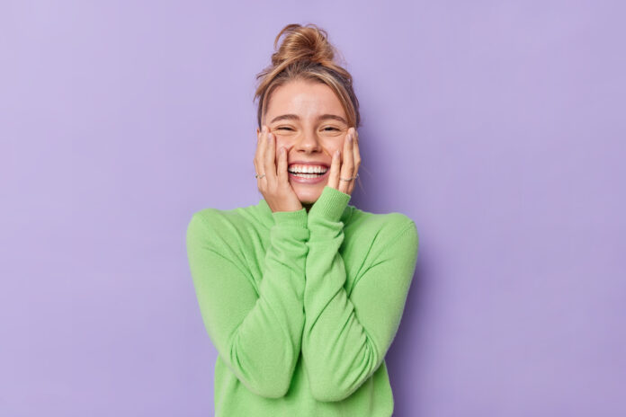 Portrait of happy young female model keeps hands on cheeks smiles broadly shows white teeth feels glad expresses sincere emotions wears green turtleneck isolated over purple studio background